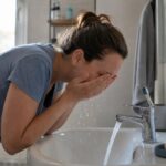 Женщина утром мягко умывает лицо чуть тёплой водой над раковиной