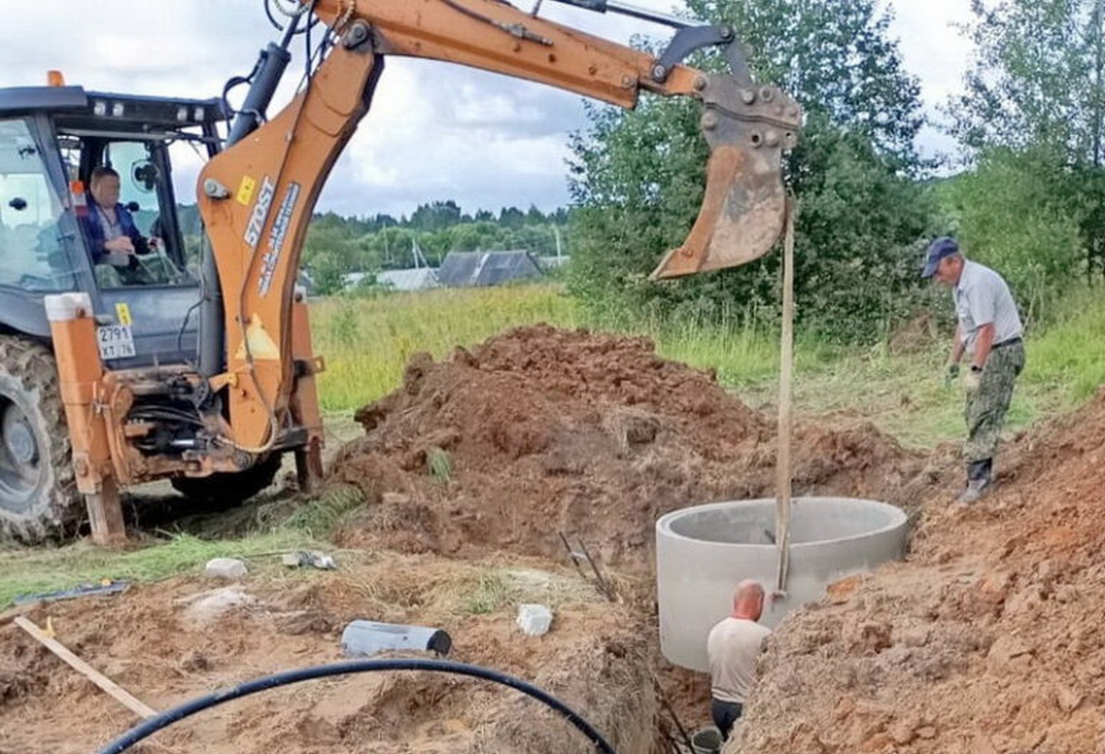 Экскаватор опускает бетонное кольцо в котлован, рядом работают двое мужчин