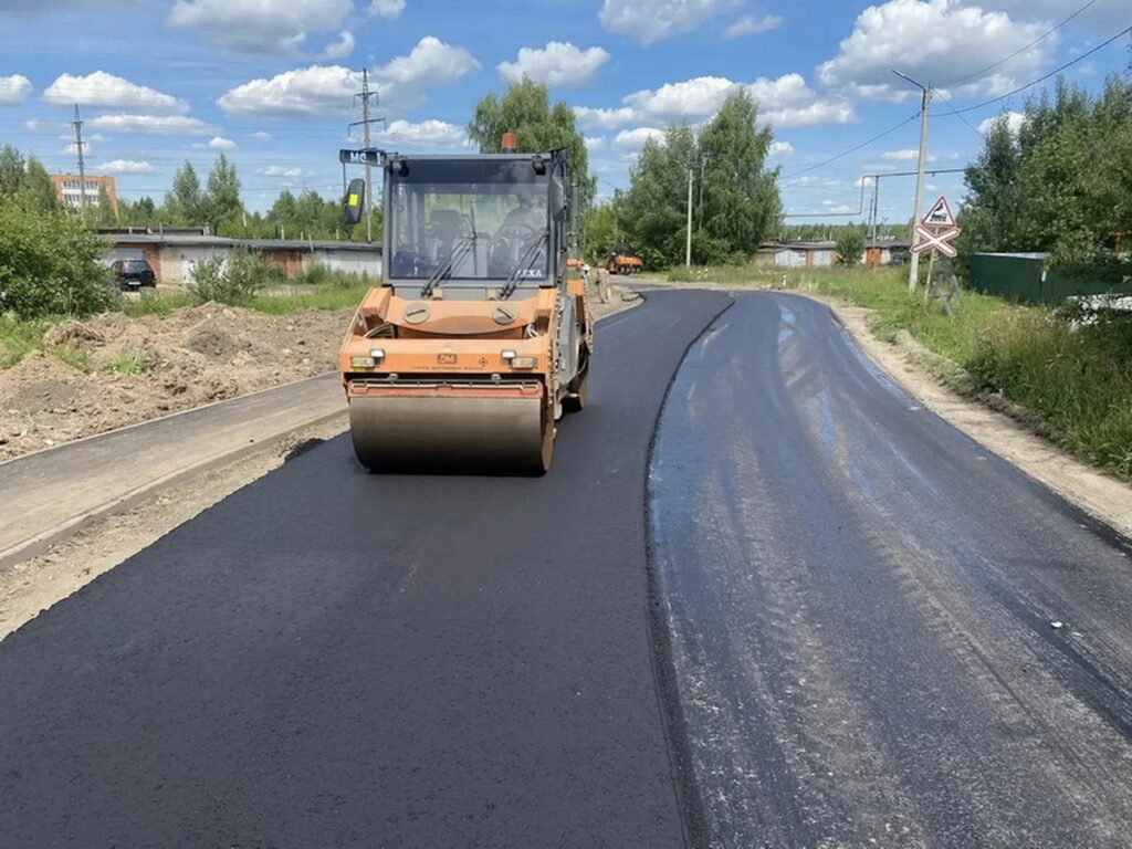 Дорожный каток укладывает свежий асфальт на изгибающейся дороге возле железнодорожного переезда