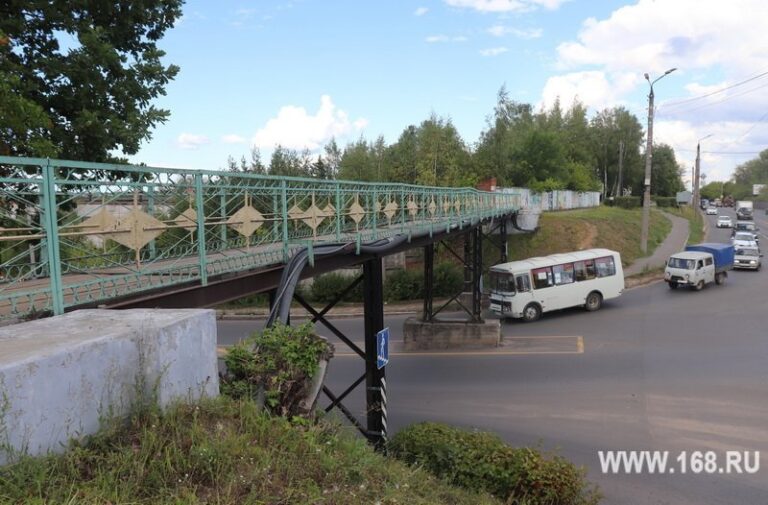 Металлический пешеходный мост с зелёными ограждениями над дорогой с автобусом и автомобилями