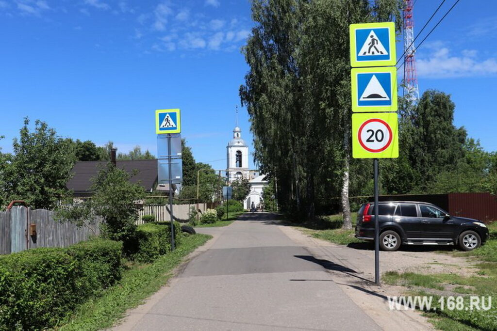 Узкая улица с дорожными знаками перехода, неровности и ограничения скорости; справа припаркован автомобиль, вдали церковь