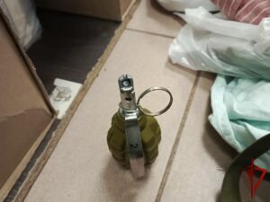 Предмет в форме гранаты с металлическим рычагом и кольцом стоит на столе среди коробок и пакетов