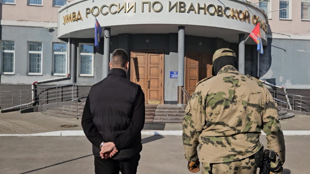 Мужчина со скованными за спиной руками и человек в камуфляже идут к входу в здание управления МВД