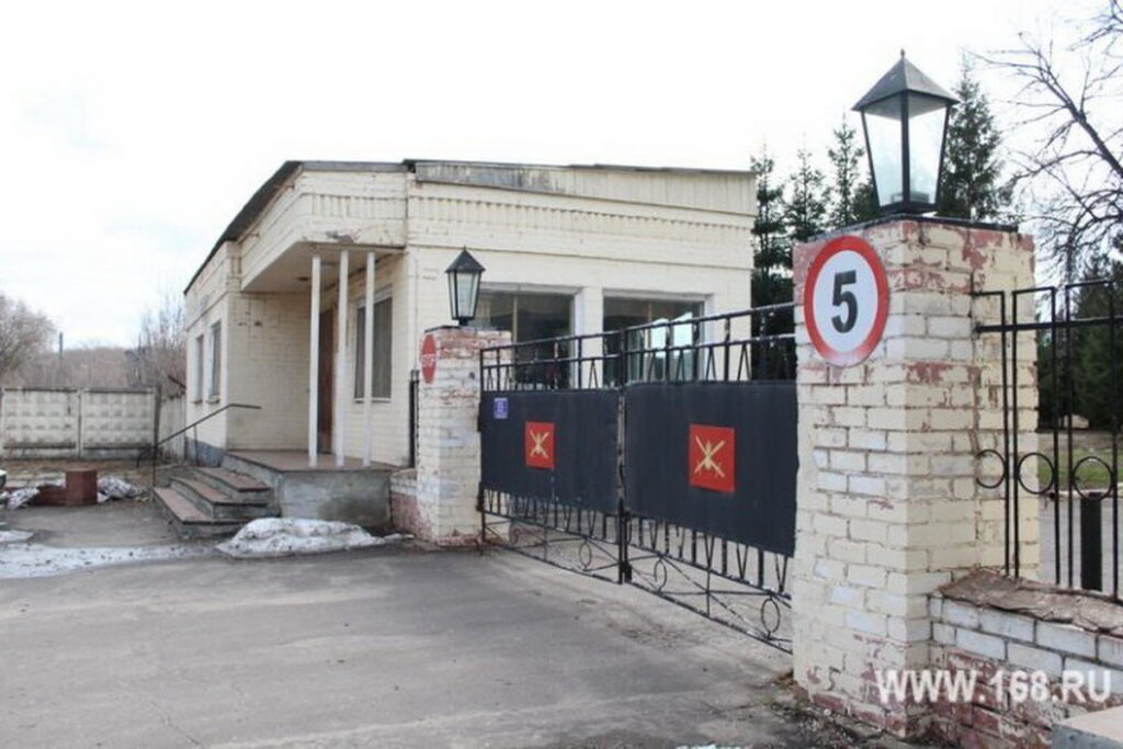 Закрытые металлические ворота между кирпичными столбами у одноэтажного здания; на столбе знак ограничения скорости 5