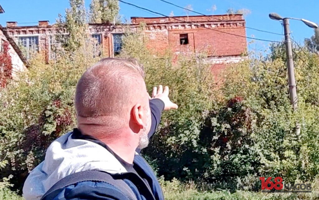 Мужчина со спины указывает рукой на заросшее краснокирпичное промышленное здание с разбитыми окнами