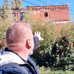Мужчина со спины указывает рукой на заросшее краснокирпичное промышленное здание с разбитыми окнами