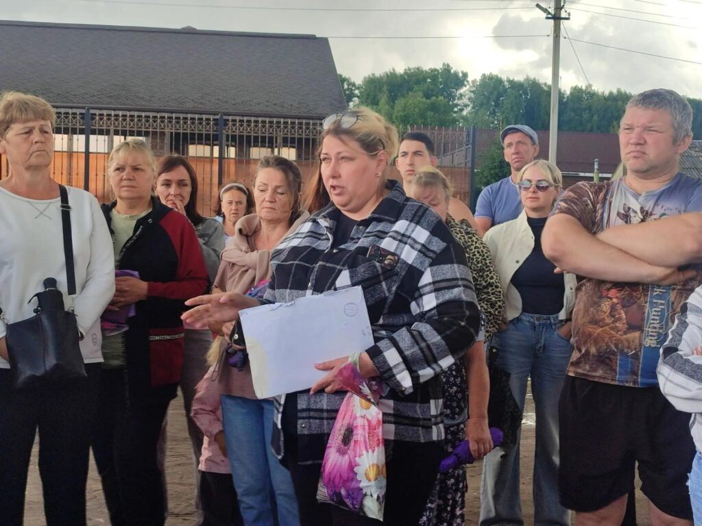 Женщина в клетчатой куртке держит бумаги и говорит перед группой людей
