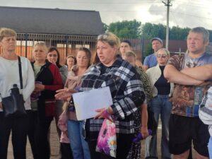Женщина в клетчатой куртке держит бумаги и говорит перед группой людей