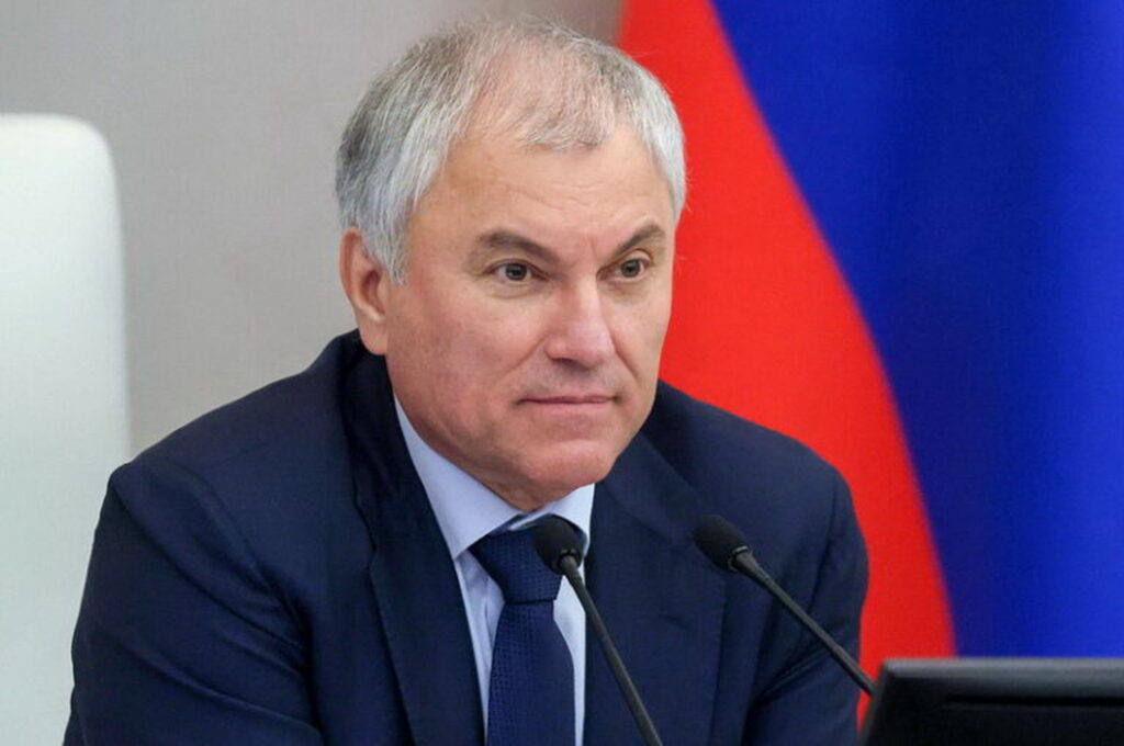 Седовласый мужчина в тёмном костюме сидит перед двумя микрофонами на фоне российского флага
