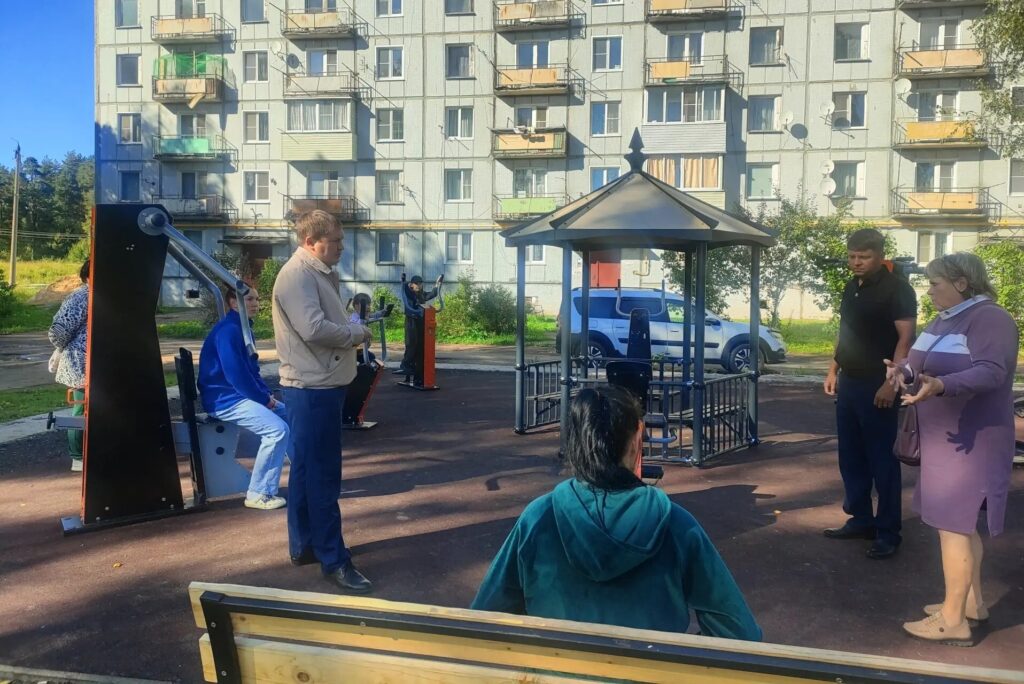 Люди осматривают площадку с уличными тренажёрами и беседкой возле многоквартирного дома