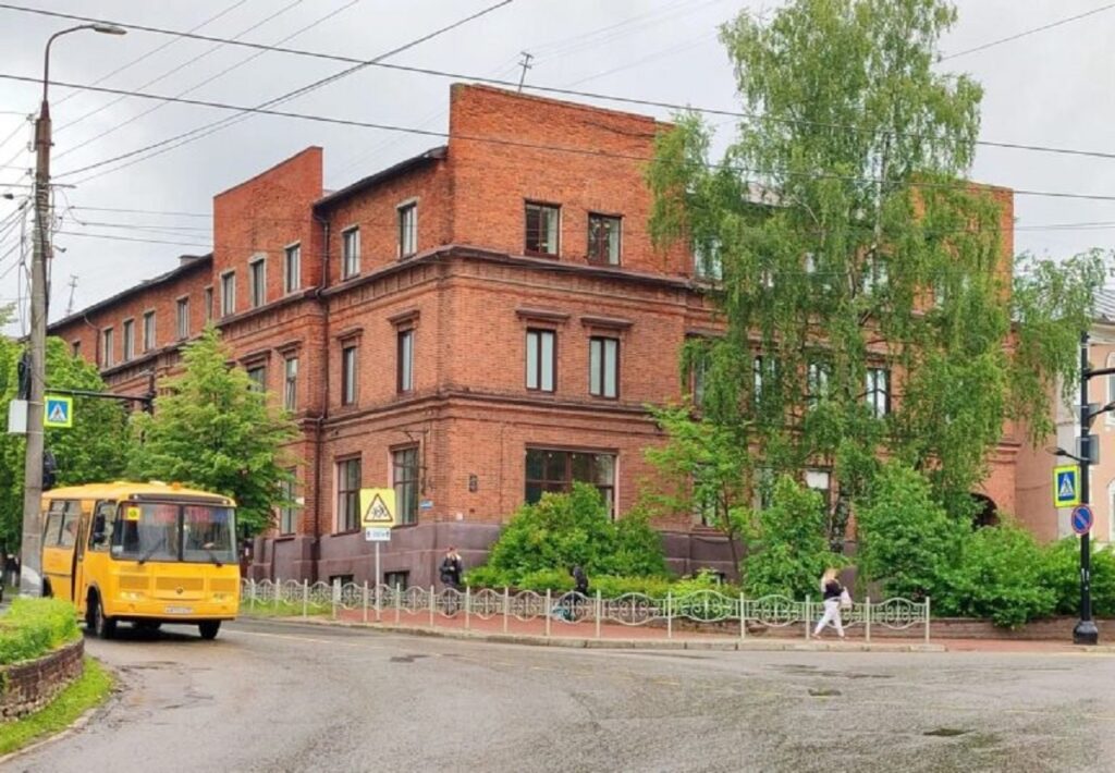 Трёхэтажное здание из красного кирпича за деревьями; по улице перед ним едет жёлтый автобус
