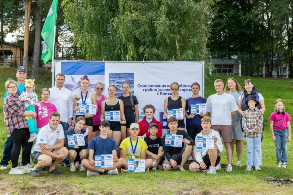 Спортсмены и болельщики позируют с грамотами и кубками на траве перед стендом соревнований