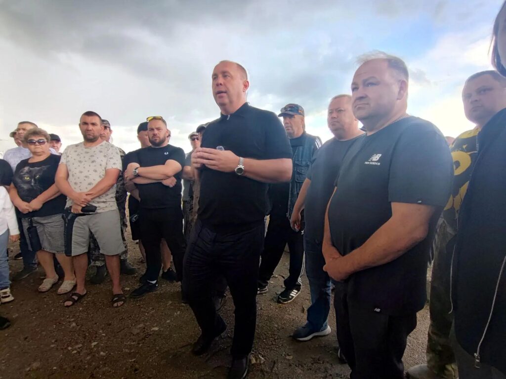 Мужчина в чёрной одежде выступает перед окружившей его группой людей под облачным небом