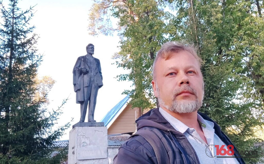 Мужчина с седой бородой фотографирует себя на фоне памятника и деревьев