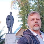 Мужчина с седой бородой фотографирует себя на фоне памятника и деревьев