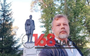 Мужчина с седой бородой фотографирует себя на фоне памятника и деревьев