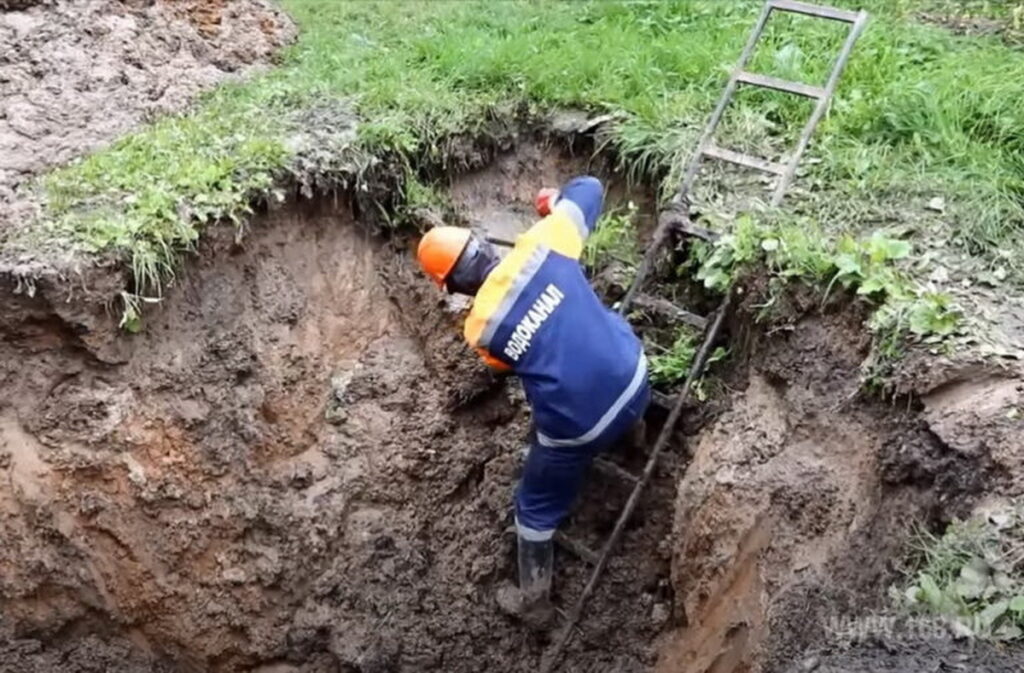 Рабочий в каске спускается по лестнице в глубокую земляную траншею