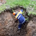 Рабочий в каске спускается по лестнице в глубокую земляную траншею