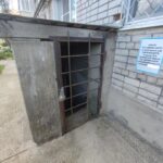 Вход в подпольное укрытие с металлической решёткой у кирпичного дома; справа на стене прикручена информационная табличка об укрытии.