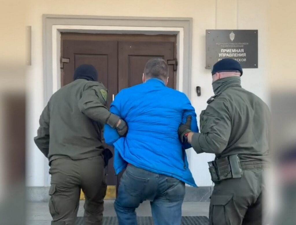 Двое мужчин в темной форме ведут мужчину в синей куртке к дверям здания