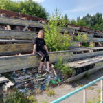 Девушка сидит на обветшавшей деревянной трибуне, заросшей молодыми деревьями; внизу разбросан мусор.