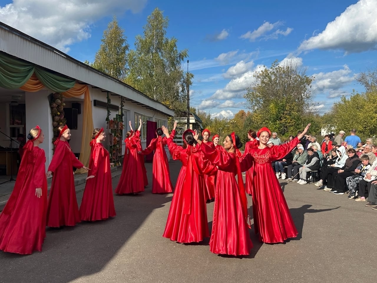 Групповое танцевальное выступление: несколько девушек в одинаковых длинных красных платьях на уличной площадке перед сидящей публикой, слева здание с декором.