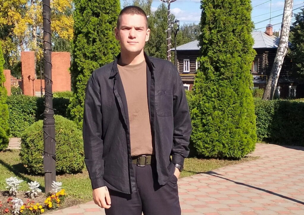 Молодой мужчина стоит на дорожке среди подстриженных кустов и деревьев, на заднем плане деревянный дом