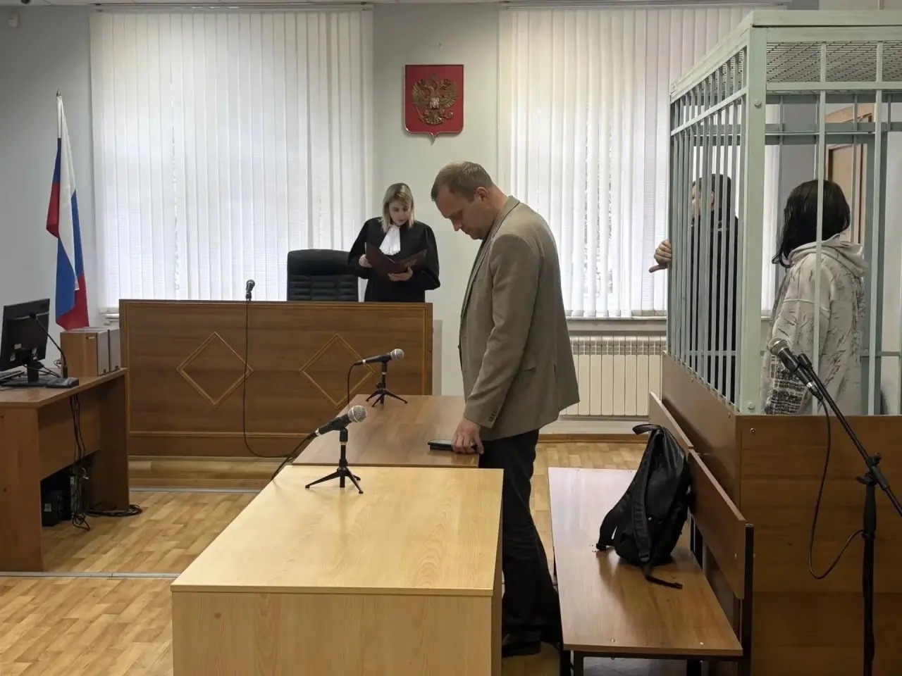 Зал суда: судья у скамьи, мужчина у стола и человек в металлической клетке справа