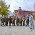 Группа военнослужащих в камуфляже и голубых беретах слушает женщину-экскурсовода на городской площади