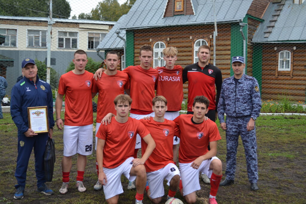 Футбольная команда в красной форме позирует с двумя мужчинами; один держит рамку с благодарностью.