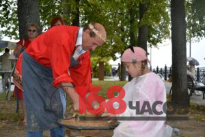 Мужчина в красной рубахе помогает девочке формовать глиняный сосуд на гончарном круге под открытым небом