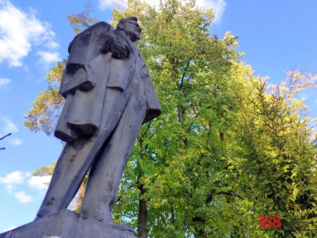 Памятник мужчине в длинном пальто, снятый снизу на фоне деревьев и голубого неба