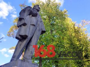 Памятник мужчине в длинном пальто, снятый снизу на фоне деревьев и голубого неба