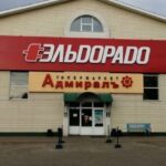 Фасад торгового центра с большой красной вывеской «Эльдорадо» и жёлтой вывеской «Адмиралъ» над входом.