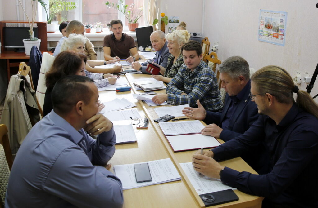 Участники заседания сидят за длинным столом с документами и обсуждают рабочие вопросы.