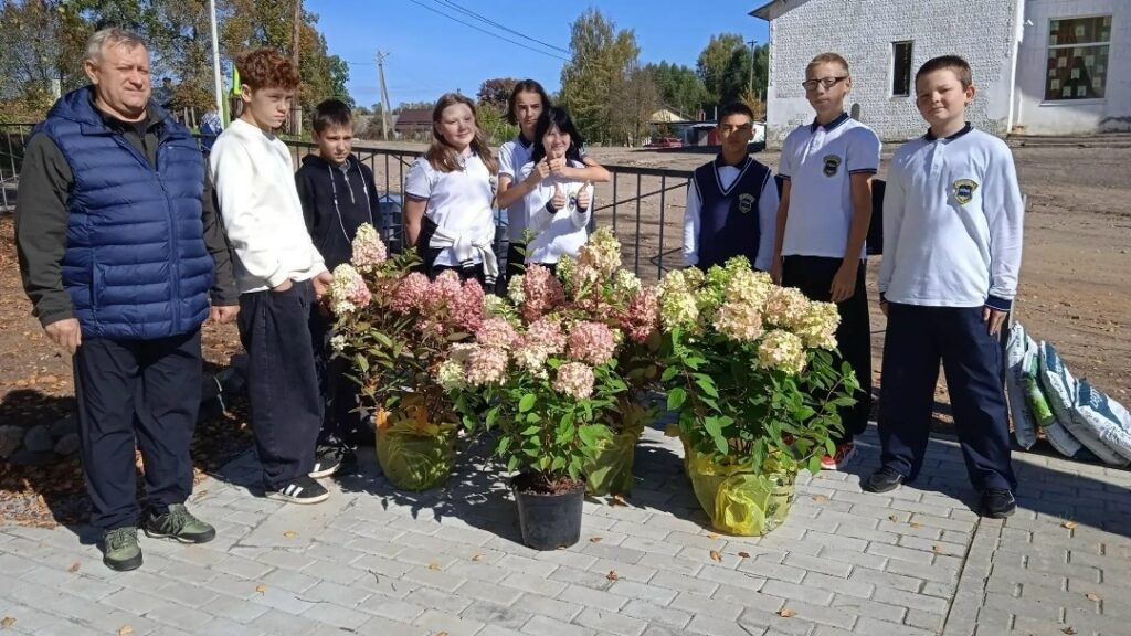 Группа школьников и взрослый стоят на площадке рядом с горшками цветущих гортензий.