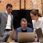 Трое коллег обсуждают работу за ноутбуком в офисе с кирпичными стенами
