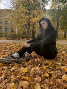 Темноволосая женщина в черной куртке сидит среди опавших листьев в осеннем сквере