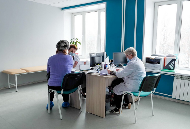 Пациентка сидит за столом в клинике напротив двух медицинских работников в белых халатах и масках; в кабинете стол с компьютерами, принтер и кушетка.