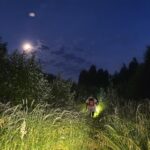 Ночная заросшая лесная просека с высоким травяным покровом; человек с рюкзаком идёт вперёд, освещая дорогу ярким светом фонаря, видна луна в небе.