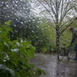 Прохладный дождливый день в нейтральном дворе: человек в лёгкой куртке проверяет припаркованный велосипед рядом с мокрой зеленью.
