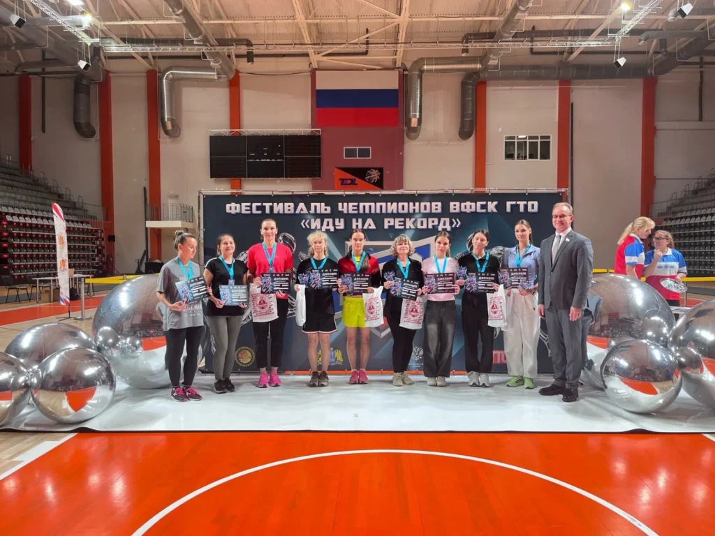 Группа спортсменок с дипломами, медалями и наградами позирует рядом с мужчиной в костюме на площадке спортивного зала