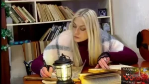 Светловолосая женщина пишет за столом рядом с фонарём и стопкой книг.