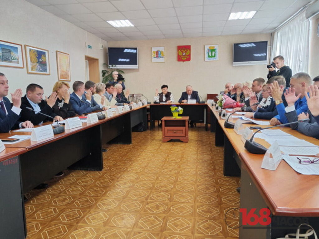Участники заседания за длинными столами поднимают руки во время голосования, двое мужчин ведут съёмку