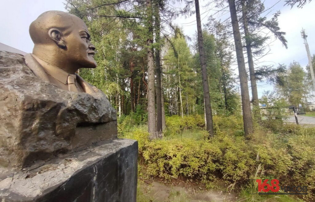 Бронзовый бюст на каменном постаменте рядом с заросшим деревьями и кустарником участком