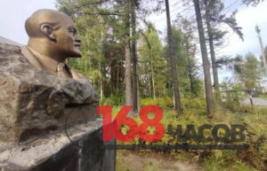 Бронзовый бюст на каменном постаменте рядом с заросшим деревьями и кустарником участком