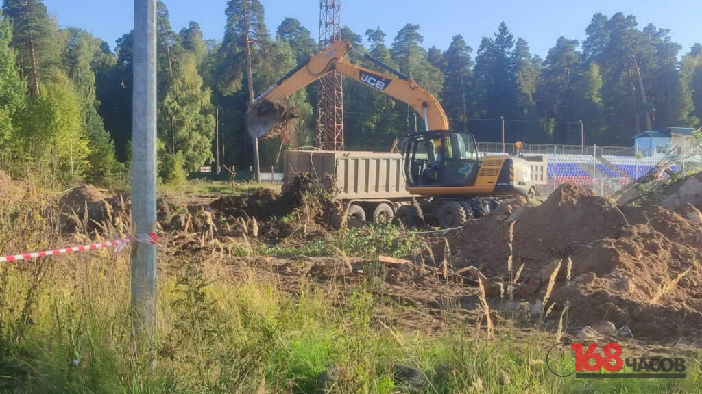 Экскаватор загружает грунт и корни в кузов самосвала на расчищаемой площадке рядом со стадионом