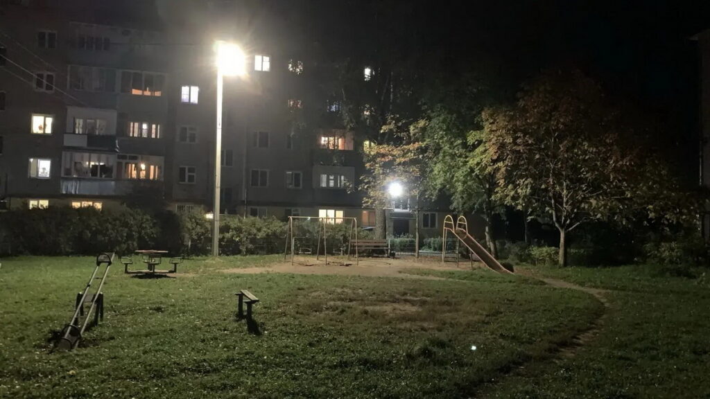 Освещённая фонарями детская площадка с качелями, горкой и лавочками во дворе многоэтажного дома ночью
