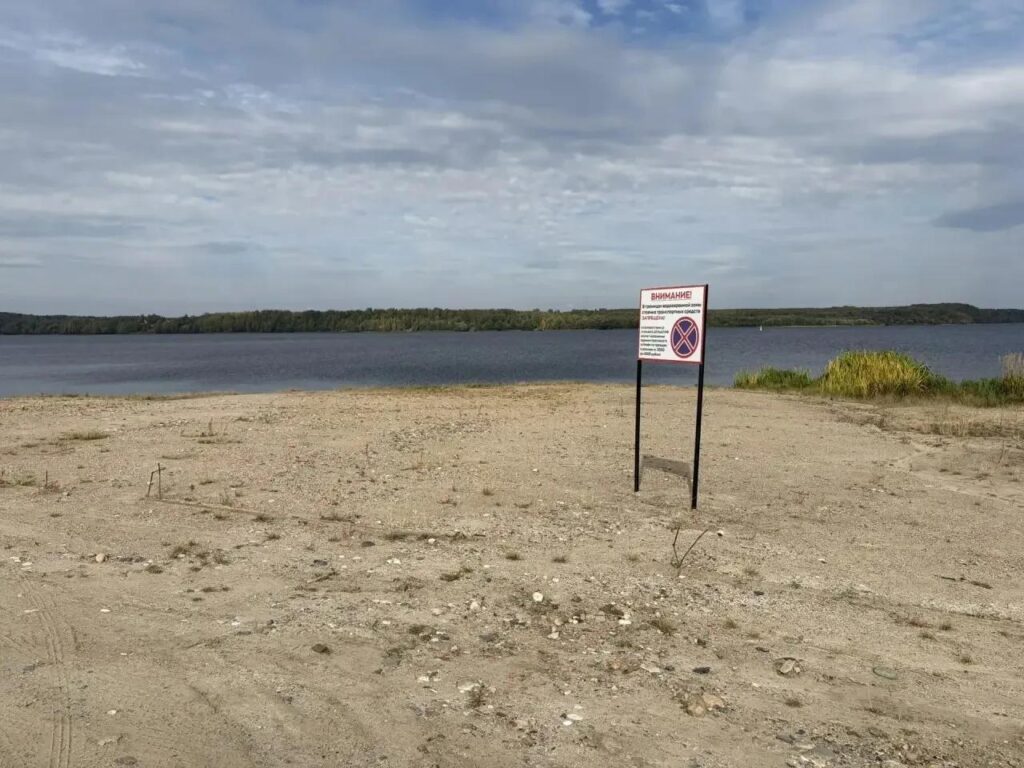 Песчаный берег водоёма со знаком о запрете движения и лесом на противоположной стороне