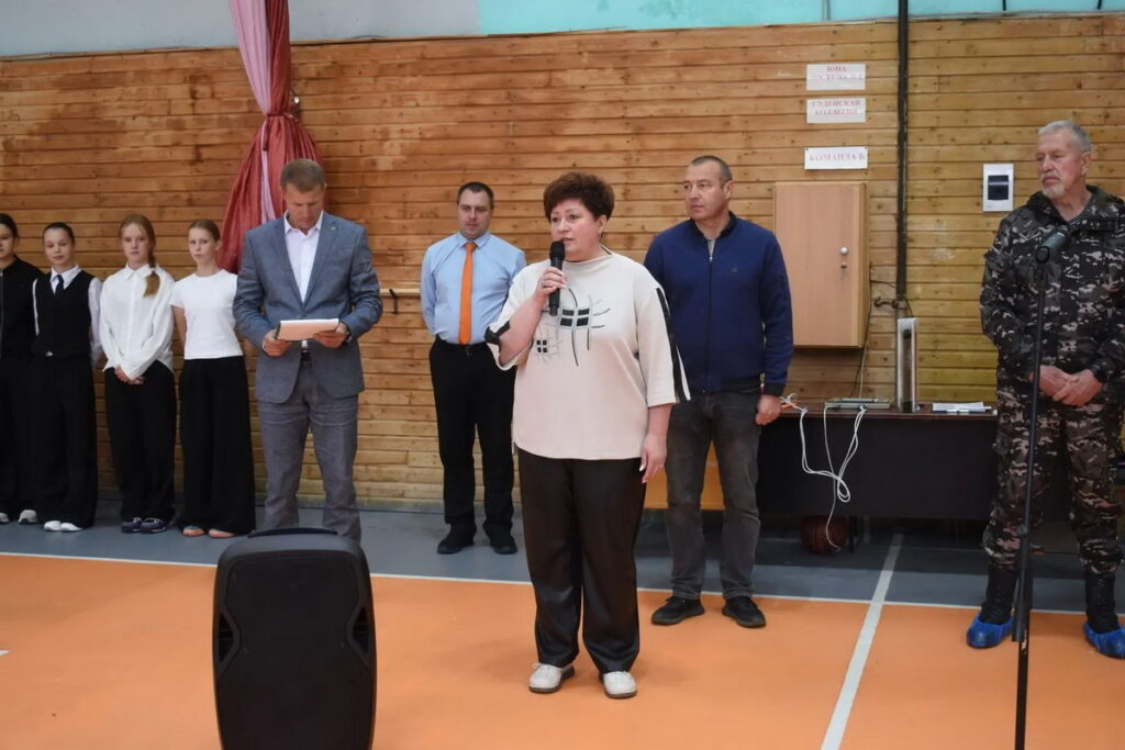 Женщина с микрофоном выступает на площадке спортивного зала перед группой взрослых и подростков